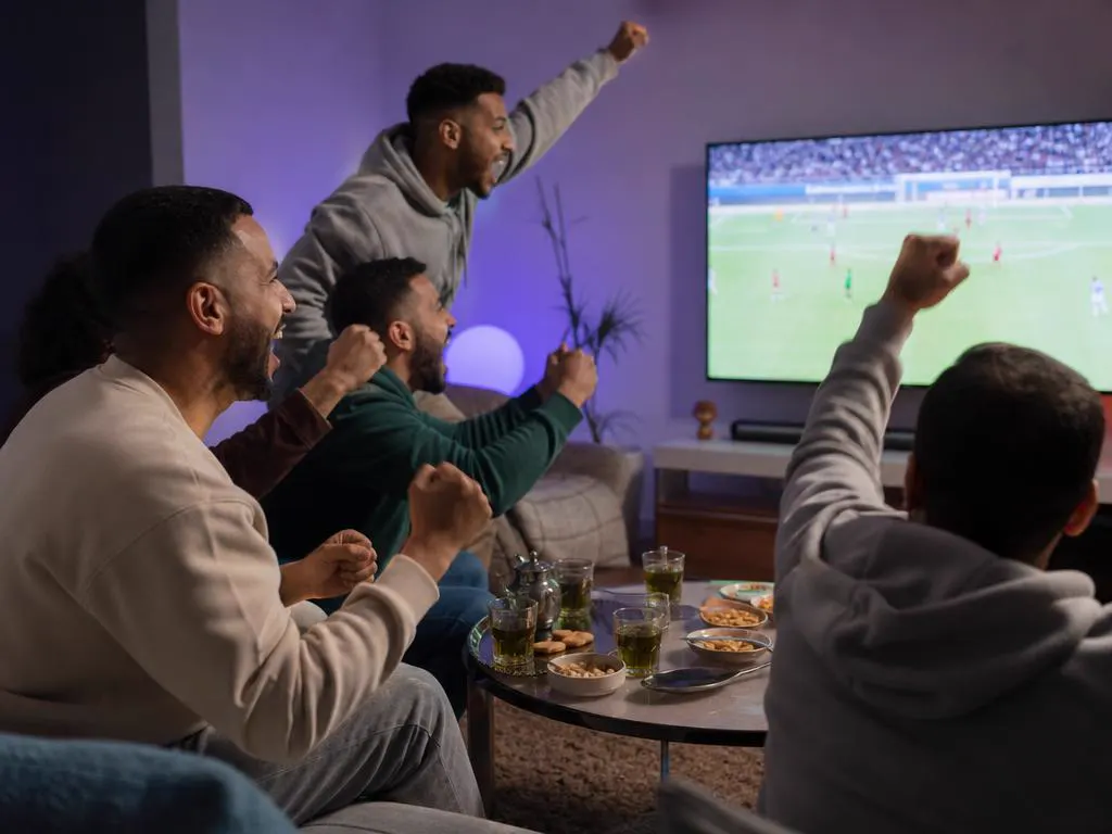 Amis dans un salon qui encouragent une équipe de football sur une grande TV avec du thé et des gâteaux