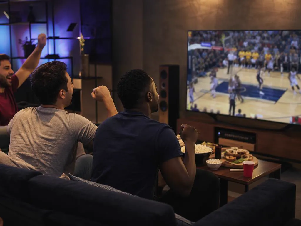 Friends watching a live basketball game on a large TV with snacks on the table