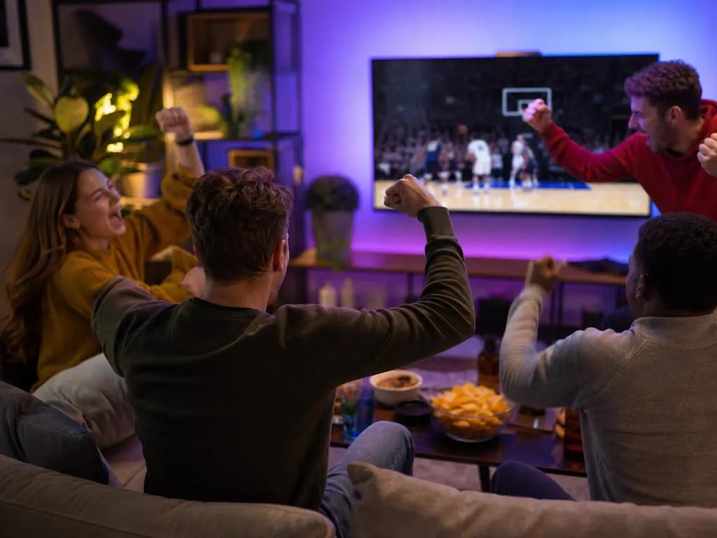 Greek friends cheering a basketball game on TV in a living room with snacks