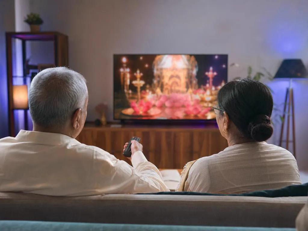 Grandparents relaxing on a sofa with a remote, watching a devotional program on TV