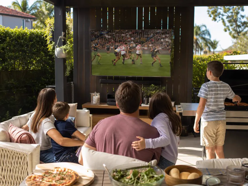 Family in a backyard barbecue setting watching a rugby league match on an outdoor TV