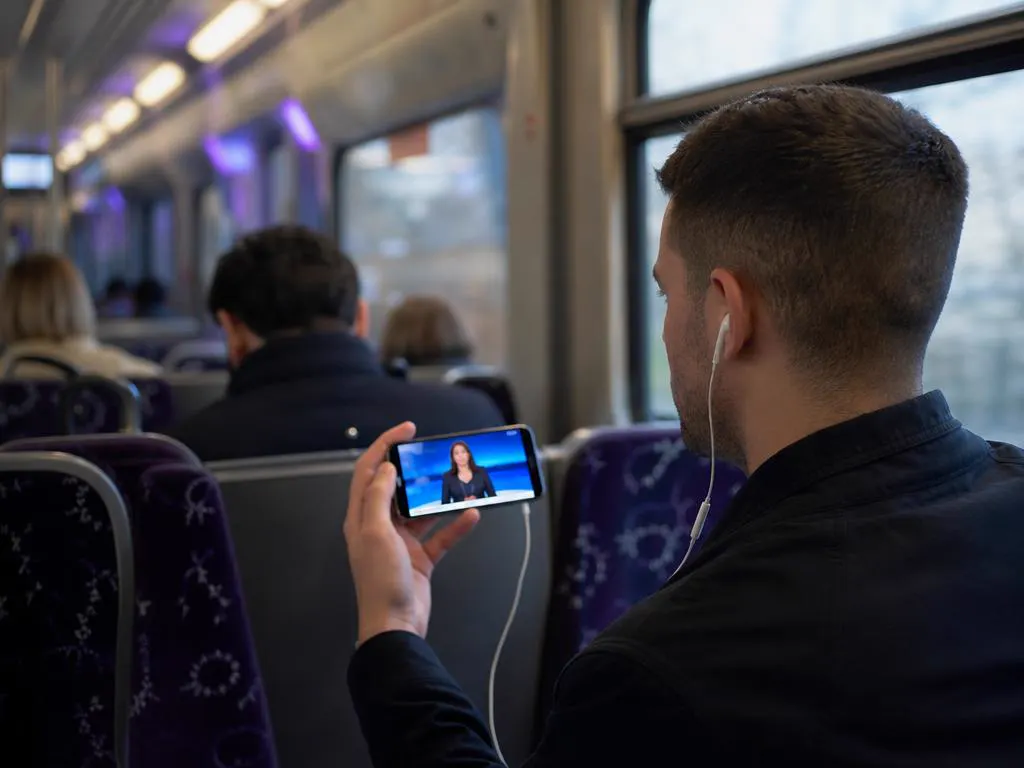 Commuter on a train watching live news on a phone with earbuds