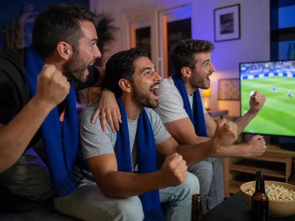 Friends in plain blue scarves celebrating a soccer goal on TV in a living room