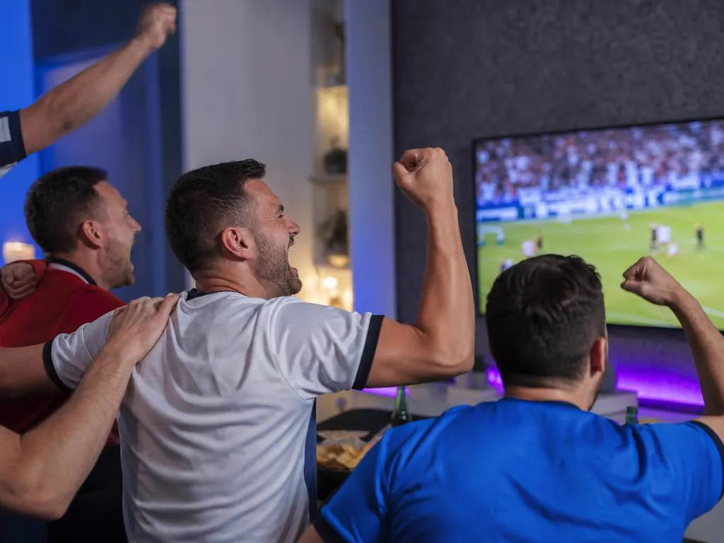 Friends in plain jerseys celebrating a goal during a soccer match on TV