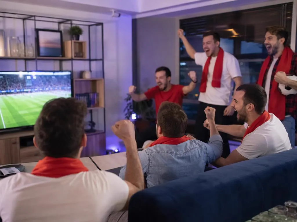 Friends in plain white and red scarves cheering a match on TV