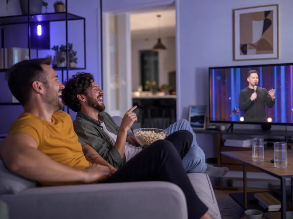 Two friends on a sofa laughing at a comedy show on a TV in a bright apartment