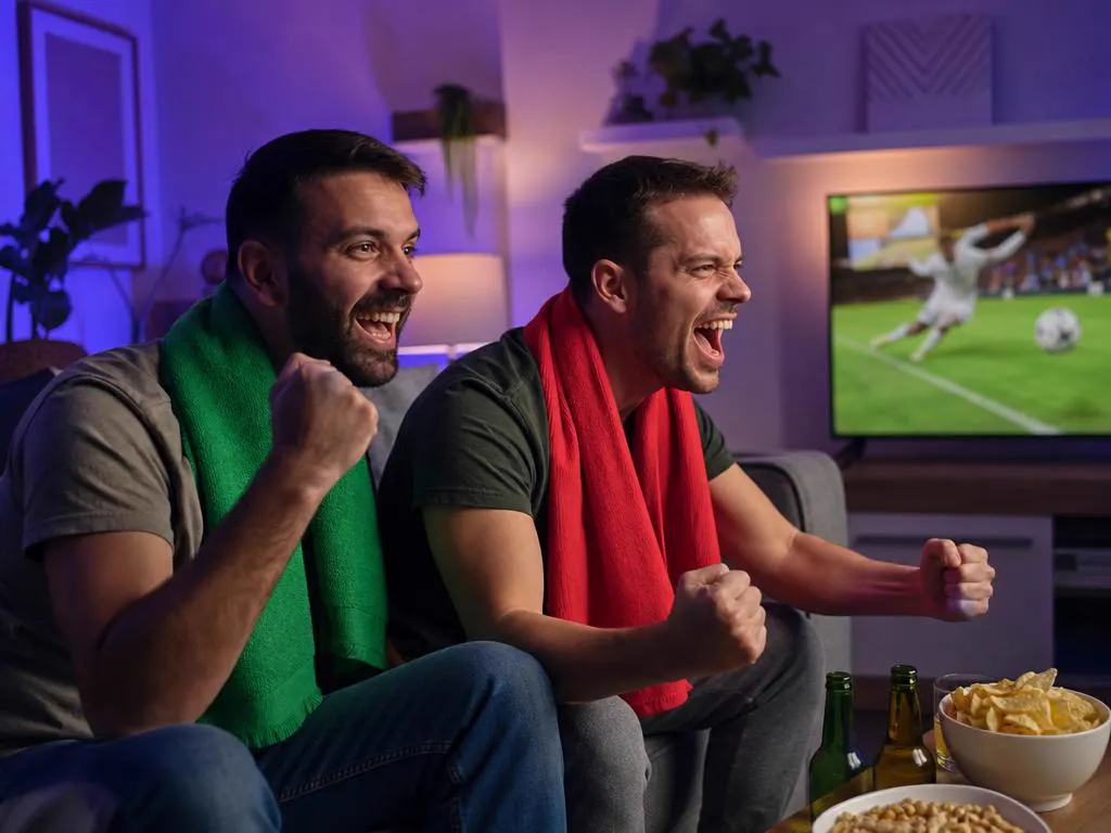 Friends in plain red and green scarves cheering a soccer match on TV